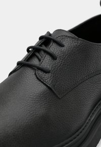 Μαύρο δερμάτινο παπούτσι με υφή, που διαθέτει στρογγυλή μύτη, λεπτές κορδόνια και μια διακριτική λεπτομέρεια κατά μήκος της σόλας.