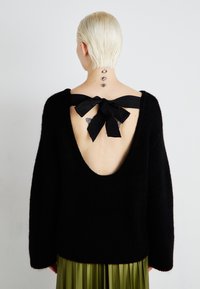 Black knitted sweater with a deep scoop back, featuring a large black ribbon tied in a bow. Model is wearing a green pleated skirt.