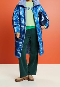 Lesklá modrá péřová bunda s kapucí, se šitým vzorem. Modelka má na sobě zelený svetr a tmavě zelené kalhoty s béžovými platformovými botami.