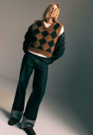Bruine en zwarte diamanten patroon wolvest over een witte shirt, gecombineerd met donkere wide-leg jeans met een rafelige zoom en bruine schoenen.