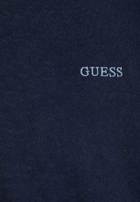 Marineblaue Stoff mit kleinem, weißen, gesticktem "GUESS"-Logo im oberen Brustbereich des Kleidungsstücks.