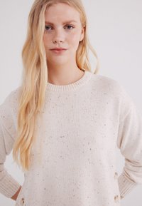 Lichtbeige trui met een ronde hals, gespetterde textuur en knoopaccenten aan de linkerkant. Gehaakt materiaal met een relaxte pasvorm.
