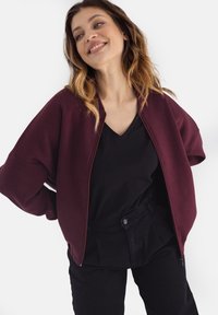 Burgundowa kurtka z zamkiem błyskawicznym o oversize'owym kroju z szerokimi rękawami, w połączeniu z czarnym topem w serek i czarnymi spodniami z wysokim stanem. Gładka tekstura.