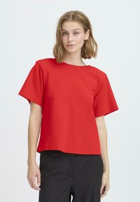 Top rojo de manga corta hecho de algodón, con cuello redondo y corte amplio y cuadrado, combinado con pantalones negros. Textura suave, diseño sencillo.