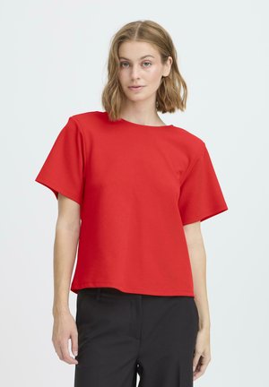 Top rojo de manga corta hecho de algodón, con cuello redondo y corte amplio y cuadrado, combinado con pantalones negros. Textura suave, diseño sencillo.