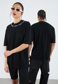 Camiseta negra de gran tamaño con bordado de alambre de pua blanco en el cuello, combinada con pantalones cortos de canalé negros y jeans negros desgastados.