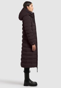 Long, puffy dark brown coat with a hood. Features zigzag stitching, front zipper, and side pockets. Paired with black ankle boots.