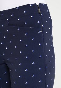 Marinblå byxor med små vita och ljusblå triangelformade mönster, med dragkedjestängning och en sidoficka med en silverknapp.