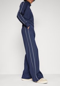 Navy tracksuit with white side stripes, featuring a zip-up jacket and wide-leg pants. Material appears soft with a smooth texture.