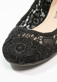 Chaussure plate noire à bout arrondi, présentant des détails complexes en dentelle florale et en tulle sur une doublure intérieure beige.