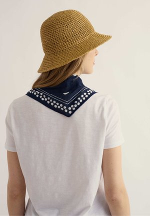 Vrouw die een beige geweven hoed en een wit shirt draagt, met een marineblauwe sjaal met witte stippen over haar schouders, van achteren gezien.