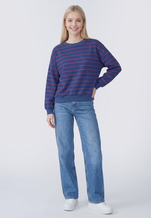 Jonge vrouw glimlacht, draagt een blauwe trui met rode strepen, lichtblauwe spijkerbroek en witte sneakers, staat ontspannen op een effen achtergrond.