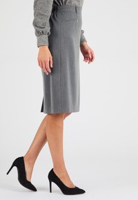 Jupe grise mi-longue dotée d'une taille froncée et d'une fermeture éclair sur le côté, assortie à des chaussures noires en daim à talons hauts. Texture lisse, coupe ajustée.