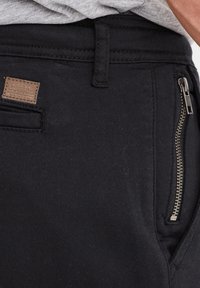 Schwarze Hose aus strukturiertem Stoff, mit einer seitlichen Reißverschlusstasche und einem kleinen braunen Leder-Label, das auf den Bund genäht ist.