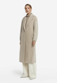 Lång, beige kappa med texturerade kabelstickade ärmar, notched krage, sidofickor och en minimalistisk design. Kombineras med en ribbad stickad tröja.