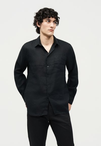 Νεαρός με καστανά σγουρά μαλλιά που φοράει μαύρο πουκάμισο με μακριά μανίκια και μαύρο παντελόνι, στέκεται με τα χέρια στις τσέπες, κοιτάζοντας πλάγια.