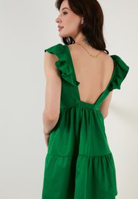 Rochie verde cu umeri volan, spate adânc în V și fustă în straturi. Fabricată dintr-un material moale, având o talie strânsă și un design fluid.