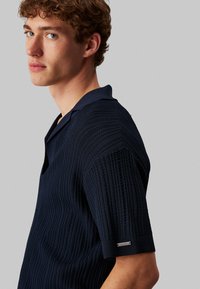 Marinblå texturerad polo tröja med V-hals och korta ärmar. Anmärkningsvärd ribbad design och varumärkesdetalj på vänster ärm.