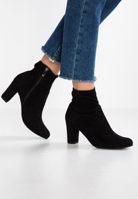 Bottes cheville en daim noir avec une coupe ajustée, détail de fermeture éclair sur le côté et un talon haut épais. Texture douce et bout pointu.