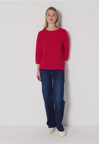 Roter Baumwollpullover mit 3/4-Ärmeln, kombiniert mit dunkelblauen Jeans und weißen Sneakers, mit einem seitlichen Kordelzug-Detail.