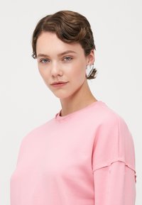 Felpa rosa chiaro con dettagli strappati e scollo a girocollo. Presenta maniche corte con aperture. Orecchini a forma di cuore argentati.