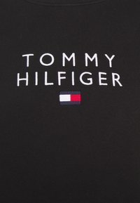 Tissu noir avec texte brodé en blanc « TOMMY HILFIGER » et un petit logo rectangulaire de drapeau avec des blocs blanc, rouge et bleu marine sous le texte.