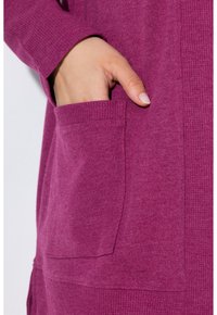 Fuchsia-Stoff mit glatter Textur, versehen mit einer Seitentasche mit geripptem Besatz. Nahaufnahme einer Hand, die in der Tasche ruht.