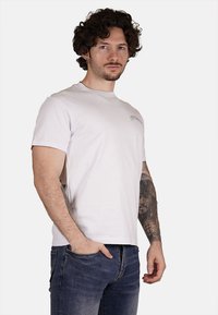 T-shirt bianca in cotone con maniche corte, collo rotondo e un piccolo logo stampato sul lato sinistro del petto. Indossata con jeans blu.