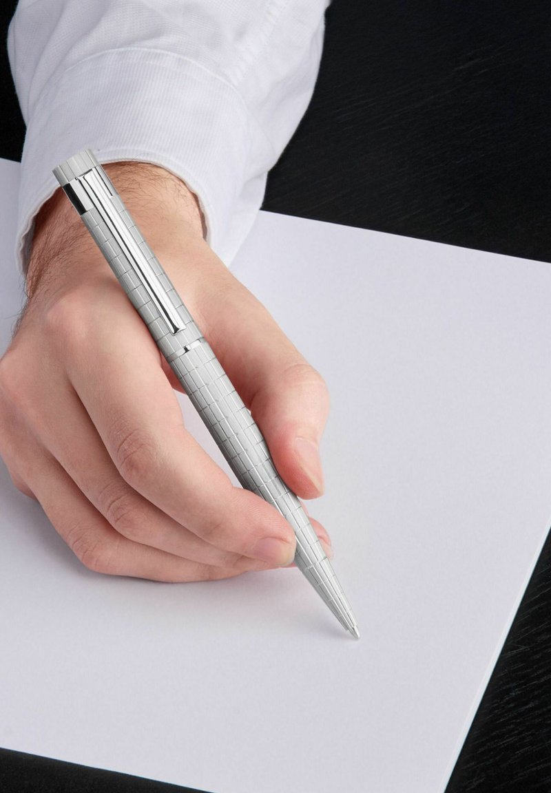 Stylo en argent avec une prise texturée tenu dans une main, suspendu au-dessus d'une feuille blanche vierge sur une surface sombre.