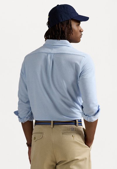 Világoskék, hosszú ujjú ing, feltűrt ujjal, khaki nadrággal párosítva, amelyet egy többszínű öv díszít. A hátsó nézet kiemeli az ing textúráját.