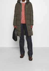 Olijfgroene pufferjas met een capuchon, gestructureerde stof, gewatteerd ontwerp; gelaagd over een roze trui, gecombineerd met donkere jeans en een zwarte tas.