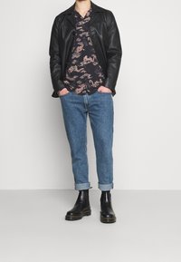 Person in schwarzer Lederjacke, dunkelblauem Shirt mit Drachenmuster, hochgekrempelten blauen Jeans und schwarzen Lederstiefeln, der gegen eine weiße Wand steht.