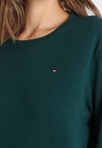 Tummanvihreä neulepusero, jossa on pyöreä pääntie. Vasemmalla puolella pieni punainen, valkoinen ja sininen logo. Pehmeä, teksturoitu kangas.