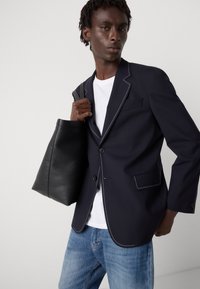 Blazer azul-marinho com costura branca, fecho de um único botão e dois bolsos. O modelo segura uma mala de couro preta e veste uma t-shirt branca e jeans azuis.