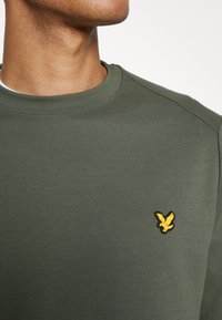 Gros plan d'une personne portant un sweat-shirt vert olive à col rond avec un petit emblème jaune d'oiseau sur le côté gauche de la poitrine.