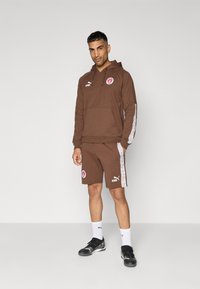 Conjunto de sudadera marrón con capucha y pantalones cortos, con detalles de logotipo, que incluye un bolsillo tipo canguro, paneles laterales blancos y zapatillas deportivas negras.