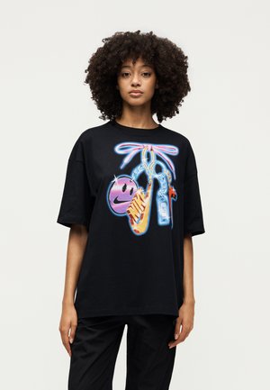 Νεαρή γυναίκα με σγουρά μαλλιά που φοράει μαύρο υπερμεγέθες T-shirt με πολύχρωμα νεον-στυλ σχέδια, όπως ένα χαμογελαστό πρόσωπο, το λογότυπο της Nike και σφυρίχτρα.