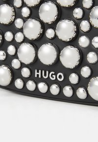 Svart tilbehør med teksturert overflate prydet med metalliske sølvnagler i varierende størrelser; har fremtredende "HUGO" merkevare under naglene.