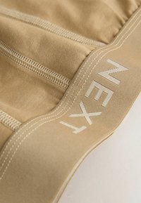 Tissu beige avec des coutures apparentes et une large ceinture élastique comportant le nom de la marque "NEXT" en texte clair.