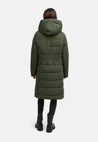 Donkergroen gewatteerde lange jas met een getailleerde taille, grote capuchon en gestoffeerde textuur. Zwarte enkelboots en leggings maken de look compleet.