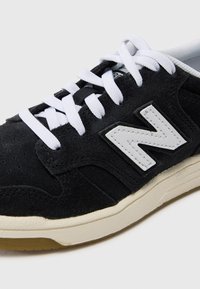 Svarte semskede sneakers med hvite snørebånd, med en markant grå New Balance-logo på siden og en beige gummisåle.