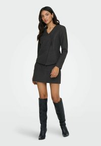 Šedé vlněné sako se zavinovacím designem, jediným knoflíkem a dlouhými rukávy, doplněné o odpovídající minisukni. Černé kozačky nad kolena.