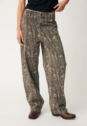 Persoon draagt losse camouflagebroek met knoop en ritssluiting, gecombineerd met donkerbruine schoenen met een vierkante neus.