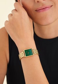Orologio quadrato color oro con quadrante verde, caratterizzato da indicatori delle ore e un bracciale in metallo intrecciato, indossato su un polso. Fondo nero.