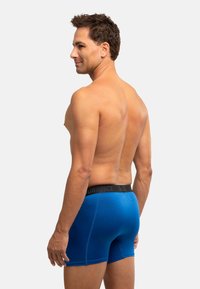 Modré boxerky s černým elastickým pasem, s hladkou texturou a přiléhavým střihem, zobrazené na muži viděném z boku.