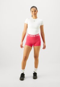 Fehér, illeszkedő, rövid ujjú felső rózsaszín Nike Pro rövidnadrággal, széles, elasztikus derékpánttal. A modellen fekete sportcipő és fehér zokni van.