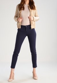 Béžová bomber bunda s volánkovými detaily, světlý top a tmavé námořnické kalhoty. Spárováno s nahými lodičkami na vysokém podpatku, celkově hladká textura.