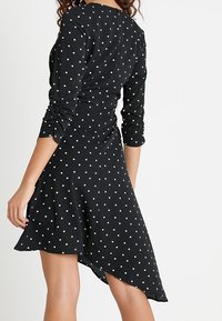 Černé šaty s puntíky, které mají dlouhé rukávy, kulatý výstřih a asymetrický lem. Vyrobené z hladké látky s jemným leskem.