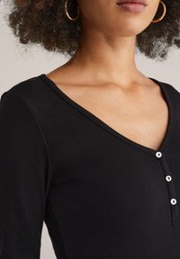 Schwarzes, geripptes Langarmshirt mit V-Ausschnitt, das drei weiße Knopfakzente an der Knopfleiste aufweist. Glatte Textur mit körpernaher Passform.