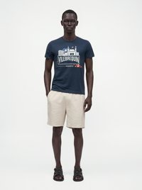 Άνδρας φορώντας μπλε σκούρο graphic t-shirt Vilebrequin Monaco, μπεζ σορτς και μαύρα σανδάλια, στέκεται με τα χέρια στις τσέπες σε λευκό φόντο.